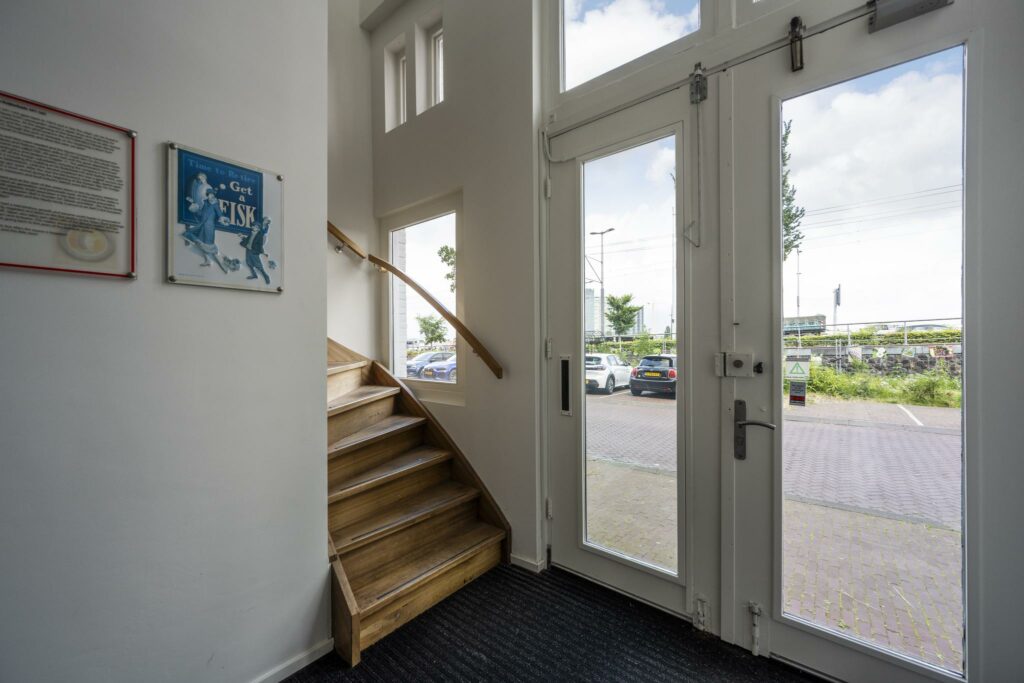 Entreehal met houten trap, glazen toegangsdeur en uitzicht op parkeerplaats aan de De Ruijterkade.